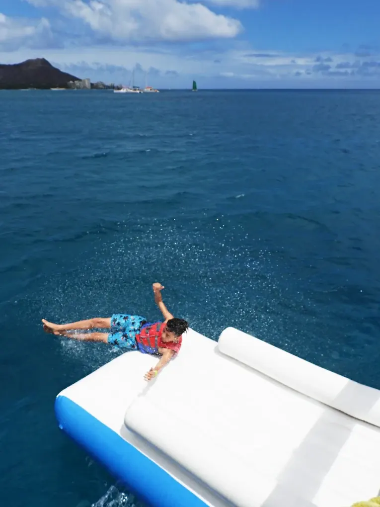 Child in a life vest sliding off the boat’s waterslide into the ocean, with Diamond Head in the distance