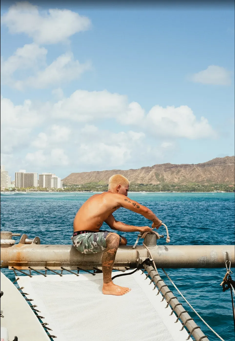 Crew member adjusting a rope on the catamaran’s beam with Waikiki hotels and Diamond Head behind